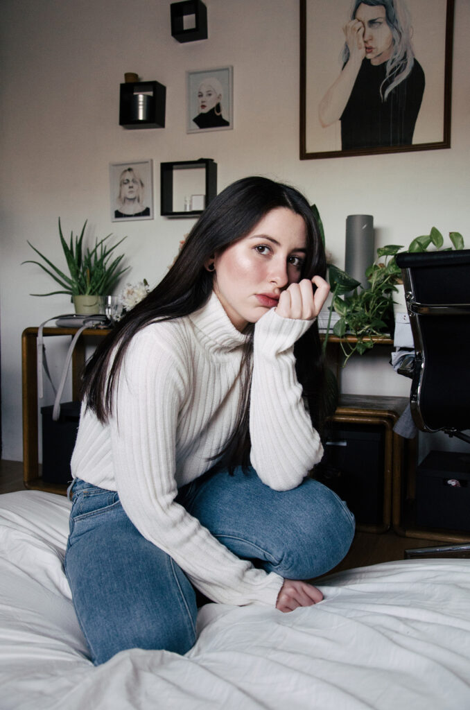 Gabriela Inzunza sits on a bed wearing a white turtleneck and blue jeans with a contemplative expression. Behind her a room is adorned with framed portraits and decorative items.
